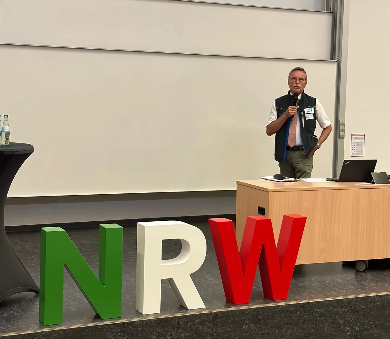 Eine Wissenschaftlerin und ein Wissenschaftler arbeiten hinter einer Glasfassade und mischen Chemikalien mit Großgeräten.